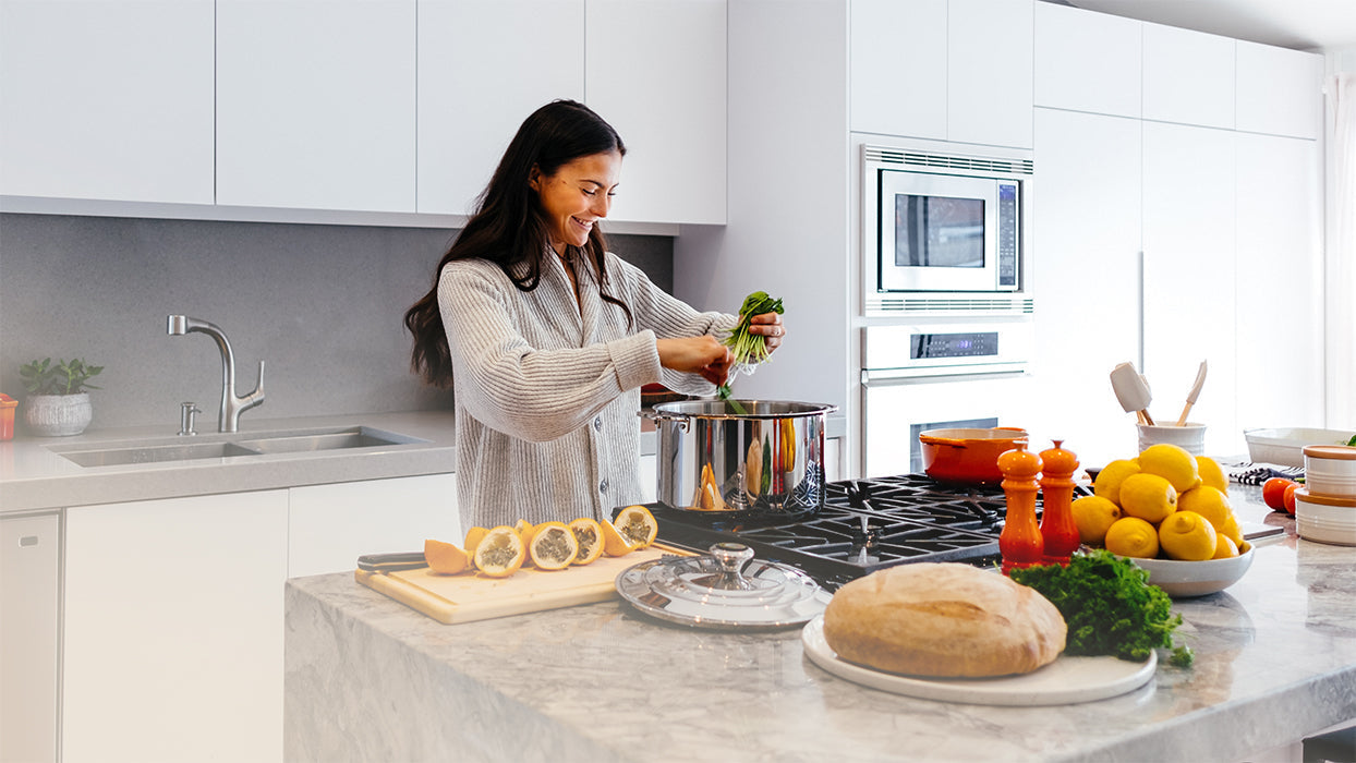 Dunkelhaarige Frau kocht in Kueche Kochinsel Brot Krauter Obst Gasherd